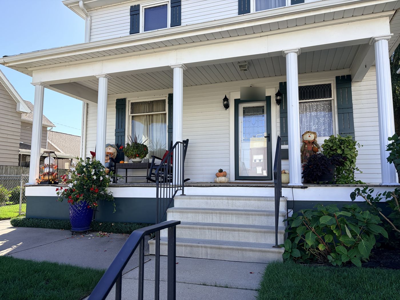 Kathy's front porch decorated for fall with white columns, green shutters, a rocking chair, flowers, scarecrows and pumpkins