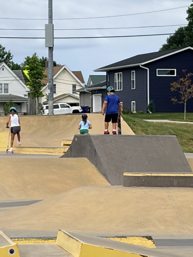 A wide view of a concrete skate park with ramps and yellow rails, a few young skaters far in the distance