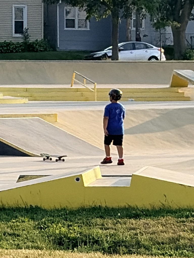 A young skater in a helmet, seen from behind, looking out over a concrete skate park with yellow ledges