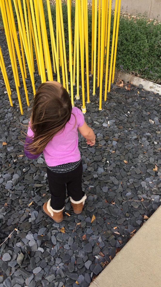 A toddler seen from behind exploring a bed of dark stones beneath tall yellow poles
