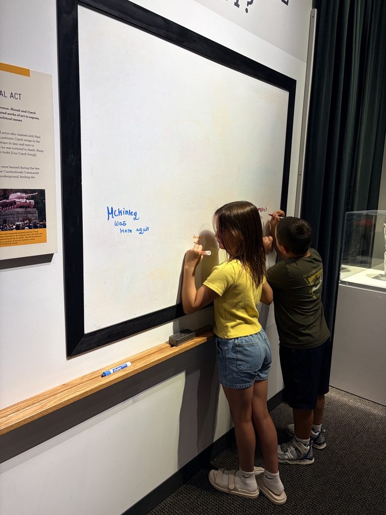 A girl and a boy writing on a large whiteboard at a museum exhibit